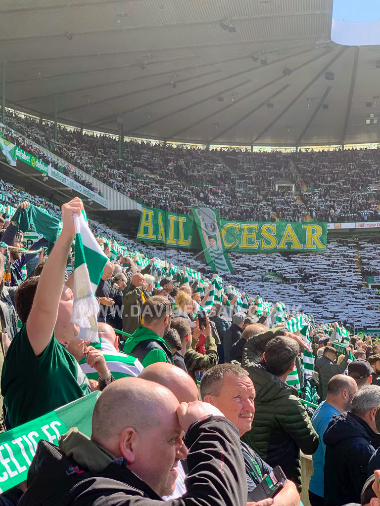 A packed stadium stand with a Hail Cesar banner and green and white scarves