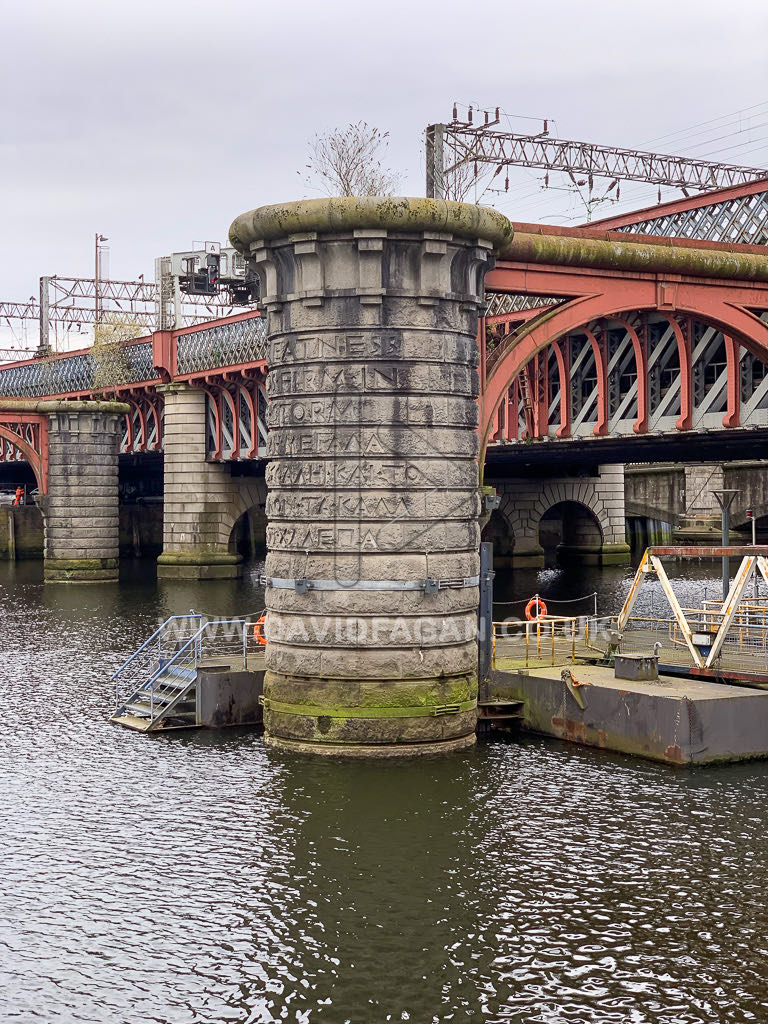 The stone piers and red ironwork of a railway bridge seen from the riverbank